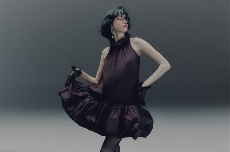 A woman in a dark purple dress and black gloves strikes a pose against a gray backdrop, showcasing high-fashion apparel in a studio setting.