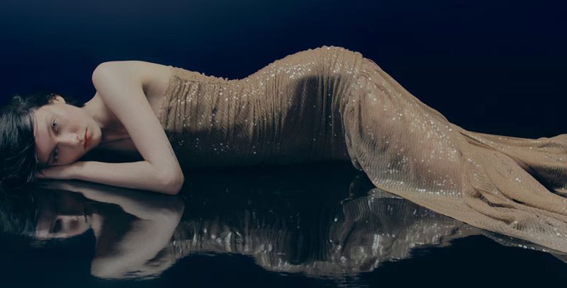 Elegant woman in a shimmering gold dress lies on a reflective surface against a dark blue background in a studio fashion portrait.