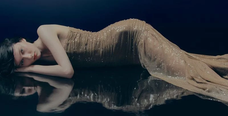 Elegant woman in a shimmering gold dress lies on a reflective surface against a dark blue background in a studio fashion portrait.