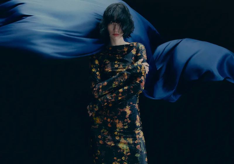 Woman in a floral print dress poses in front of a draped blue silk fabric and a black background in a fashion or editorial setting.