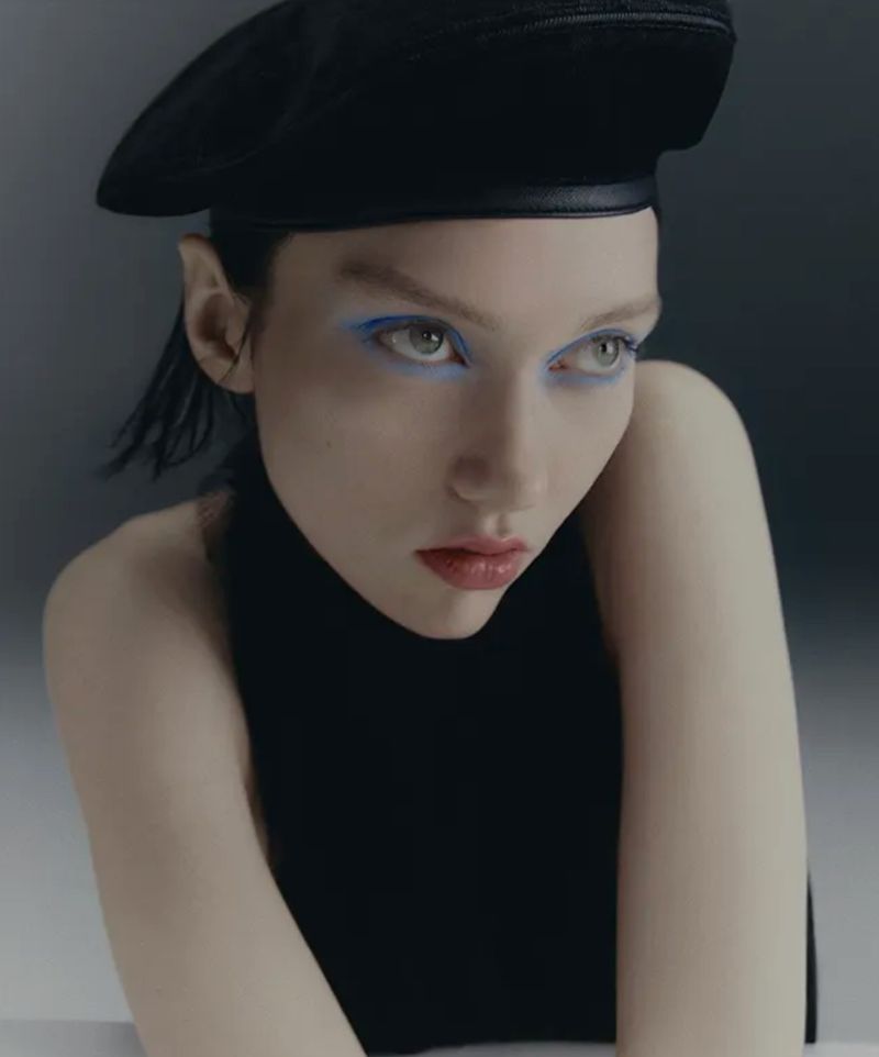 Close-up of a woman wearing a black beret and bright blue eyeliner, looking down with a slight frown against a gray background.
