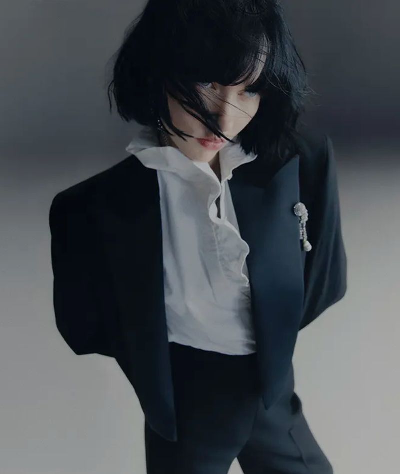 Portrait of a stylish adult woman with dark hair wearing a black tuxedo jacket and white frilled shirt in a studio setting.
