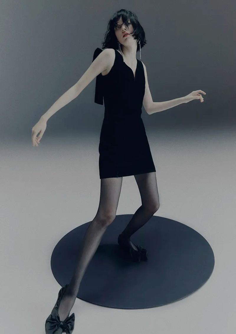 A model in a black dress and tights stands on a dark gray platform against a gray background in a studio fashion shot.