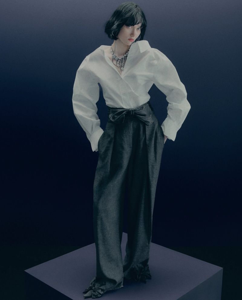 Woman posing in an oversized white shirt, wide-leg black trousers with tied waist, and a silver necklace on a gradient blue backdrop.