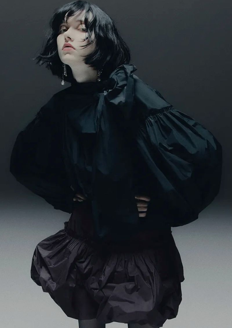 Fashion portrait of a female model wearing a voluminous black blouse and ruffled skirt, posing in a studio setting with soft, diffused lighting.