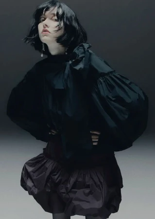 Fashion portrait of a female model wearing a voluminous black blouse and ruffled skirt, posing in a studio setting with soft, diffused lighting.