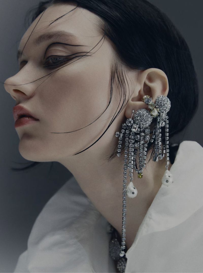 A woman with dark hair poses in a studio. She is wearing a white shirt and large crystal earrings. Her eye makeup is striking.