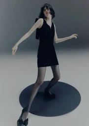 Model Steinberg in a black sleeveless mini dress, sheer black tights, and bow heels posing on a round black platform.