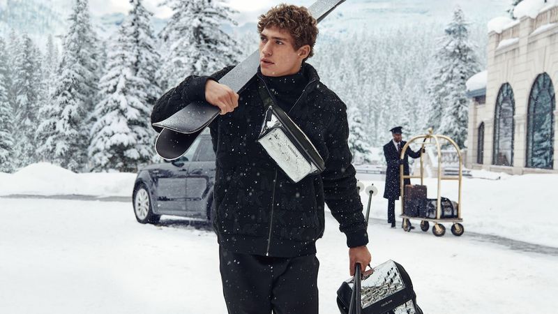 A man in winter apparel walks in a snowy landscape carrying skis, with a luxury vehicle, hotel porter, and baggage cart visible.
