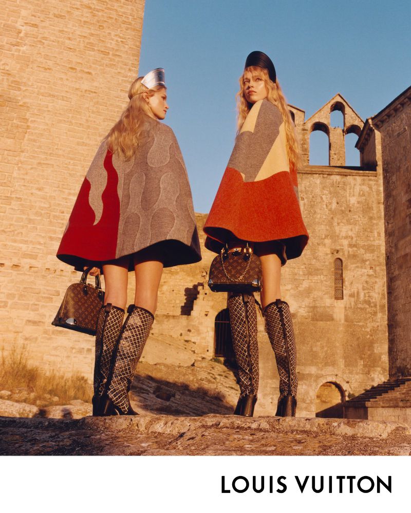 Two fashion models pose in capes and knee-high boots outside of a stone building while holding purses in an outdoor setting.
