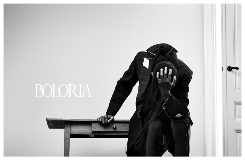 Monochrome fashion portrait of a model covering their face in a dark structured coat seated beside a wooden table for Boloria.
