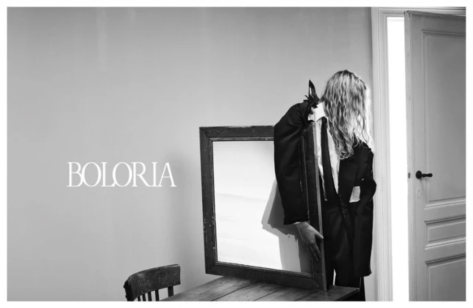 Black-and-white fashion shot of an androgynous individual in a suit holding a mirror in front of an open doorway, indoor setting.