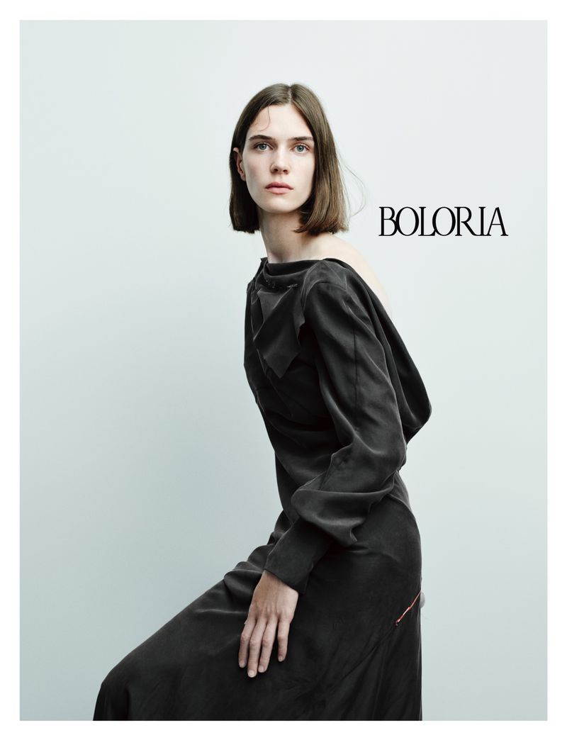 Model with a bob haircut wearing a draped black long-sleeved gown against a plain studio background.