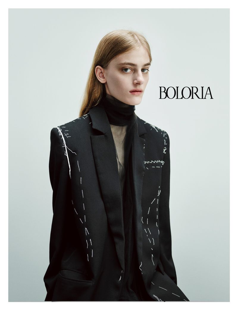 Fashion portrait of a blonde female model in a black blazer with white stitching detail against a bright, neutral backdrop.
