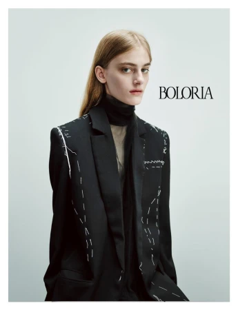 Fashion portrait of a blonde female model in a black blazer with white stitching detail against a bright, neutral backdrop.