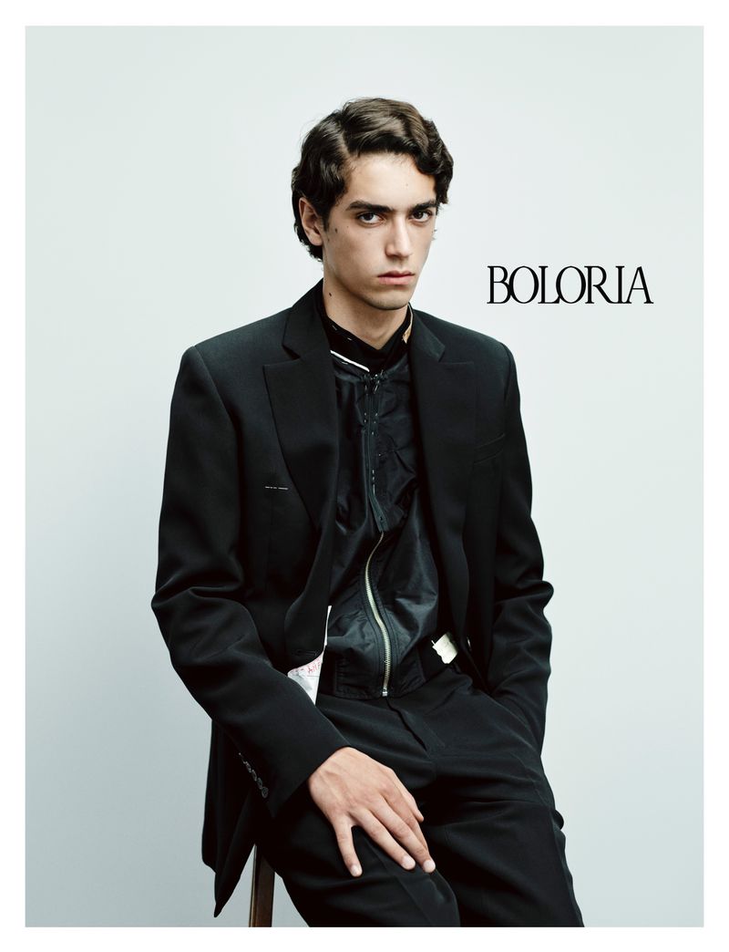 A fair-skinned male model with dark wavy hair, wearing a black suit and sitting against a grey studio backdrop in a portrait shot.