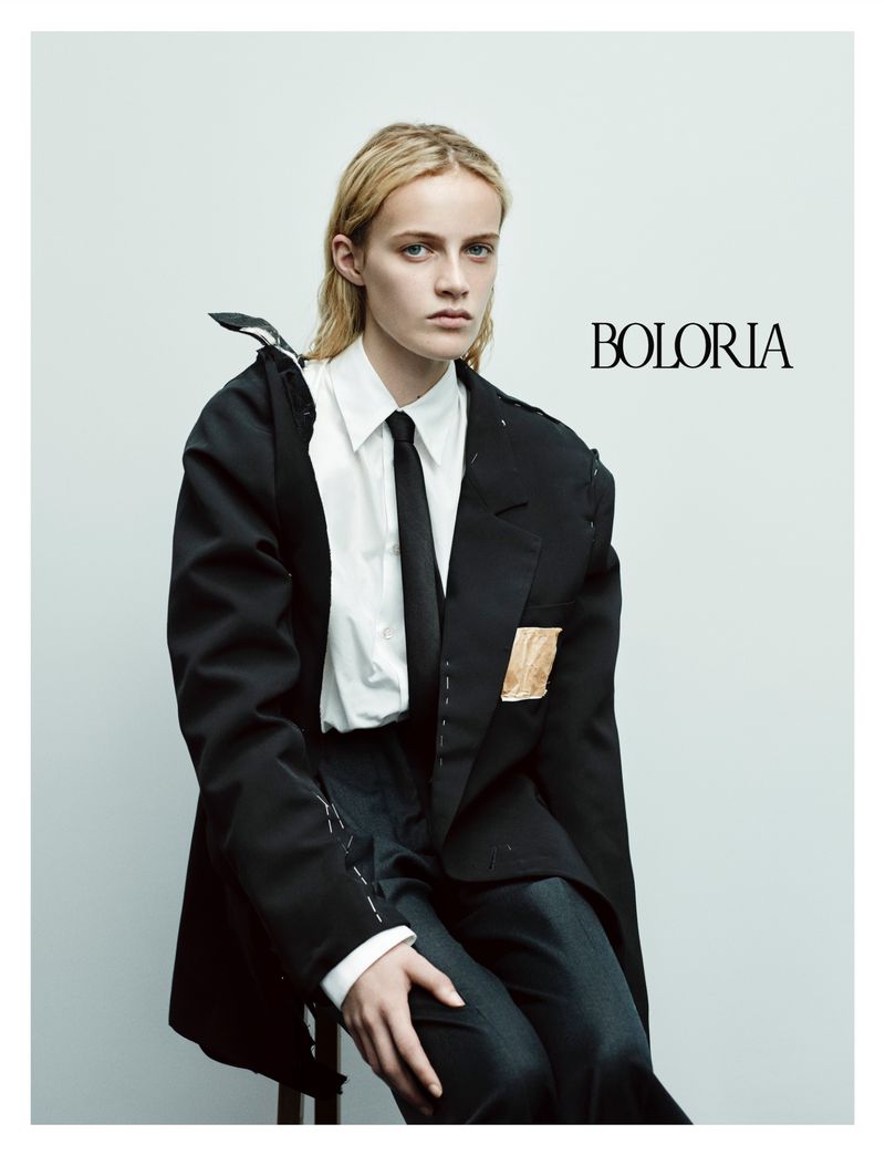 Editorial fashion shot of a slender blonde model in an oversized deconstructed black blazer, white button-down shirt, and gray slacks.