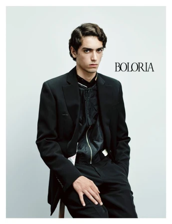 Male model seated wearing a black tailored suit jacket and zipped top against a plain studio backdrop.