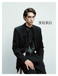 Male model seated wearing a black tailored suit jacket and zipped top against a plain studio backdrop.