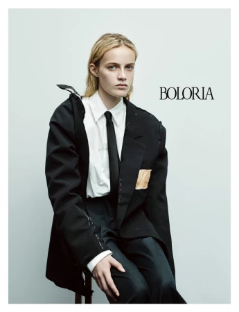 Model seated wearing a black tailored blazer, white button-down shirt, and black tie against a pale grey studio background.