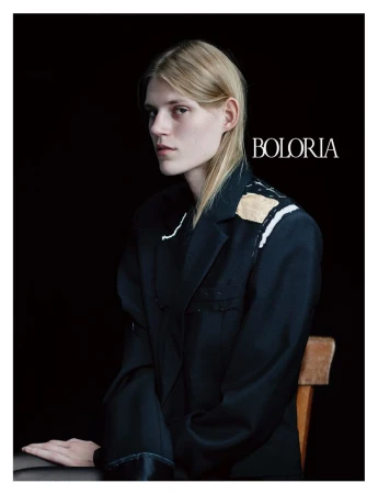 Model wearing an oversized deconstructed black blazer with white basting stitches seated against a dark seamless backdrop for Boloria.