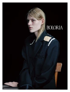 Model wearing an oversized deconstructed black blazer with white basting stitches seated against a dark seamless backdrop for Boloria.