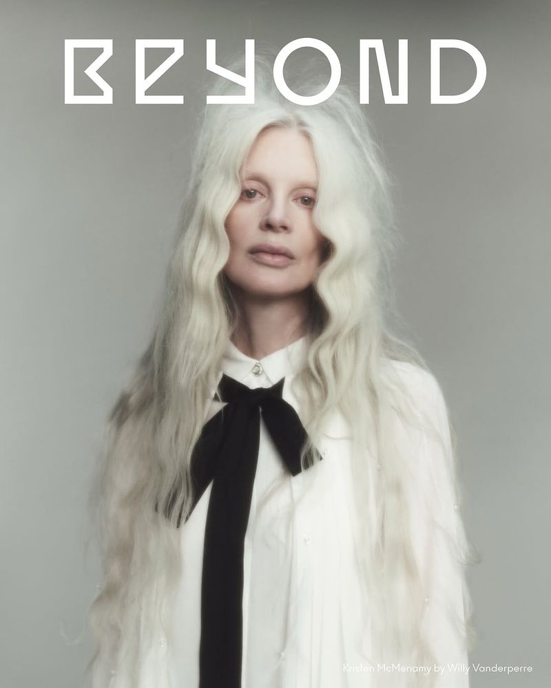 Portrait of Kristen McMenamy with flowing white hair, wearing a white blouse and black bow tie against a neutral grey backdrop.