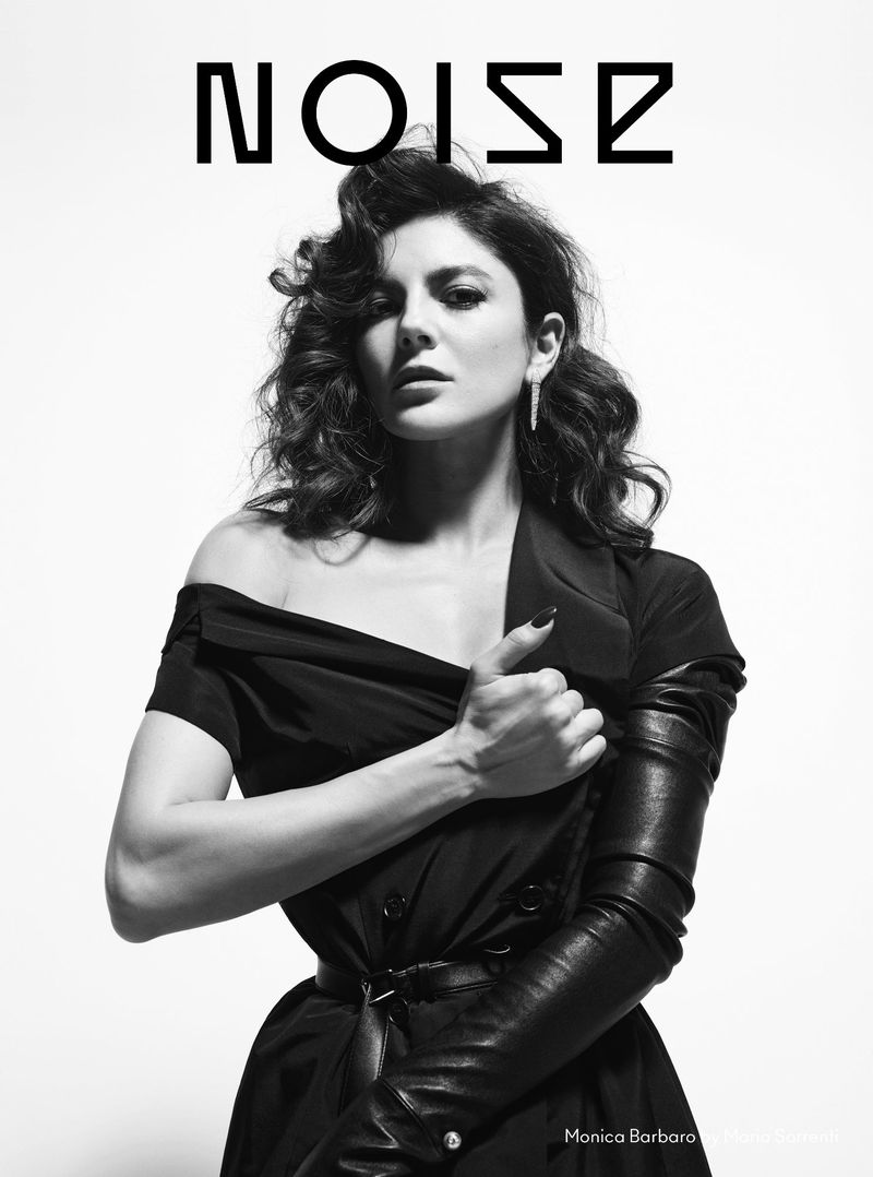 Black-and-white portrait of Monica Barbaro with dark hair and statement earrings. She wears an off-the-shoulder dress with leather gloves.