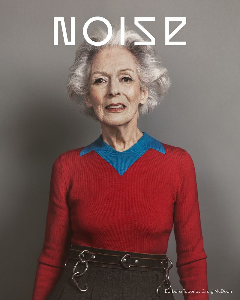 Portrait of an elderly woman with gray hair wearing a red sweater with a blue collar against a gray background.