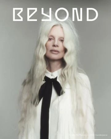 Model Kristen McMenamy with long white hair wearing a white shirt with a black ribbon tie against a grey background