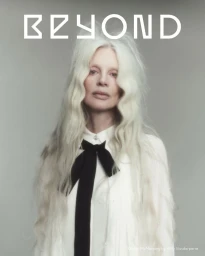 Model Kristen McMenamy with long white hair wearing a white shirt with a black ribbon tie against a grey background