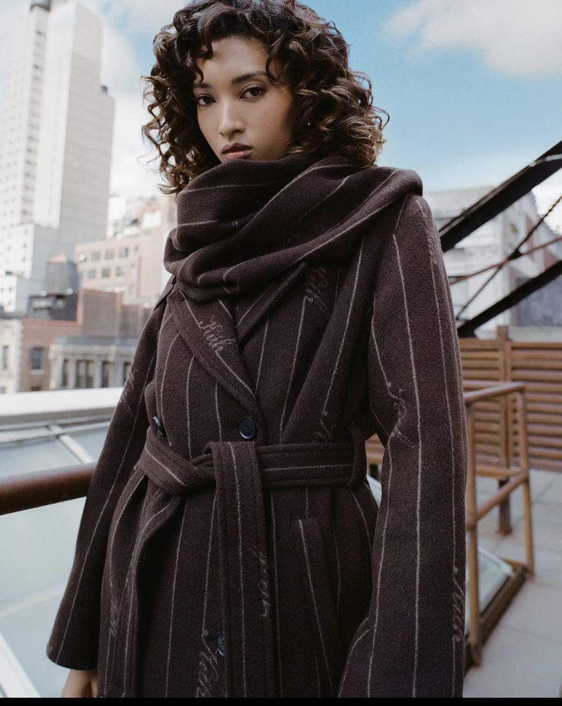 Woman with curly hair wearing a brown striped coat and scarf on a city rooftop with a cityscape in the background on a sunny day.