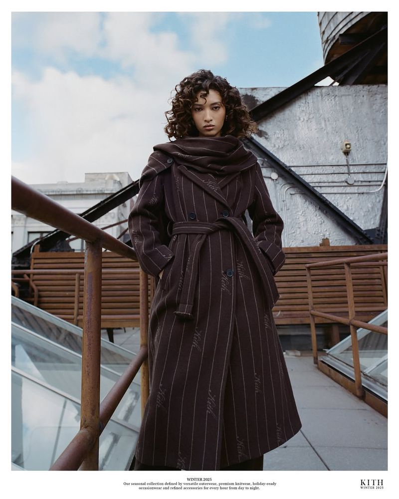 A woman wearing a brown belted coat poses on a rooftop with an urban background, showcasing a high-fashion, editorial style.