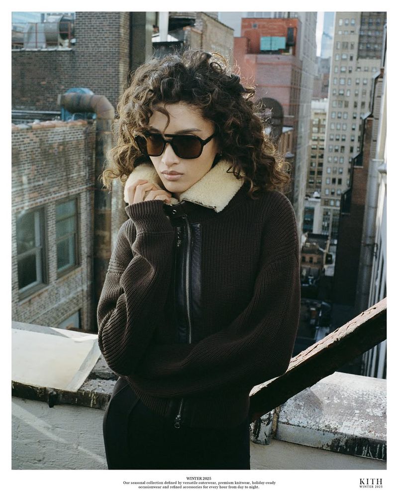 Adult female model with curly hair wears sunglasses and a brown collared jacket in an urban setting with tall buildings.