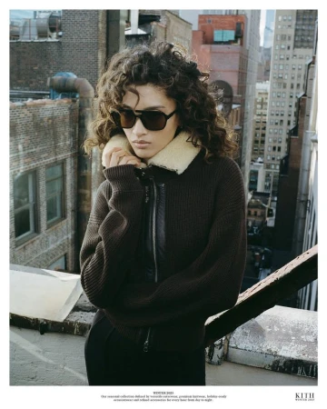 Adult female model with curly hair wears sunglasses and a brown collared jacket in an urban setting with tall buildings.