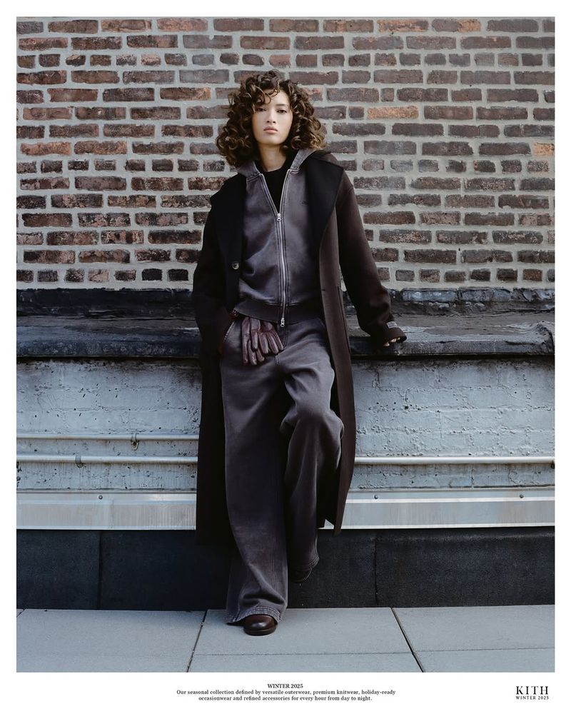 A brunette woman with curly hair poses in a grey tracksuit and long brown coat. She is leaning against a wall outdoors.