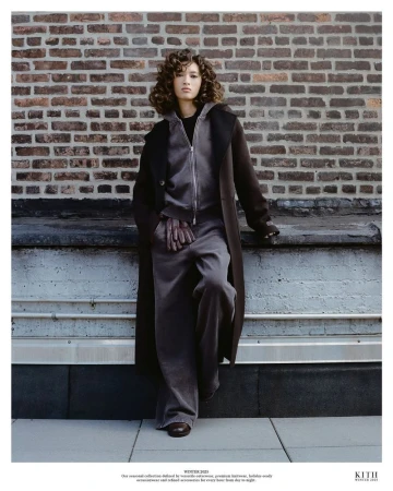 A brunette woman with curly hair poses in a grey tracksuit and long brown coat. She is leaning against a wall outdoors.