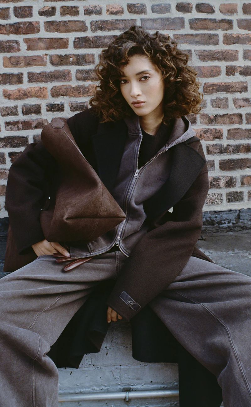 A young woman with curly hair poses outdoors in streetwear-inspired outerwear, wide-leg pants, and a tote bag in front of a brick wall.