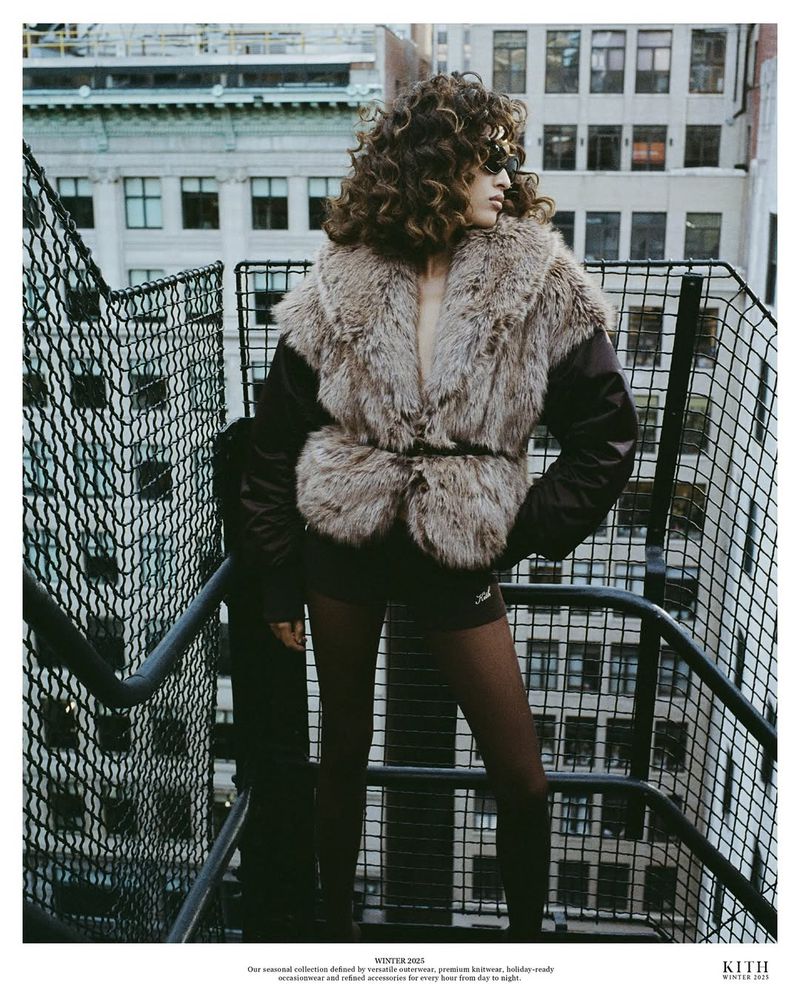 Woman poses in a fur coat on an urban rooftop with a cityscape backdrop. She wears dark tights and a jacket with brown fur.