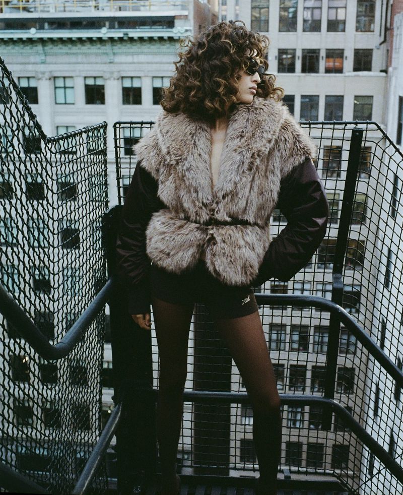 Woman in fur jacket on metal stairs in urban setting with city buildings in the background, wearing dark sunglasses and brown tights.