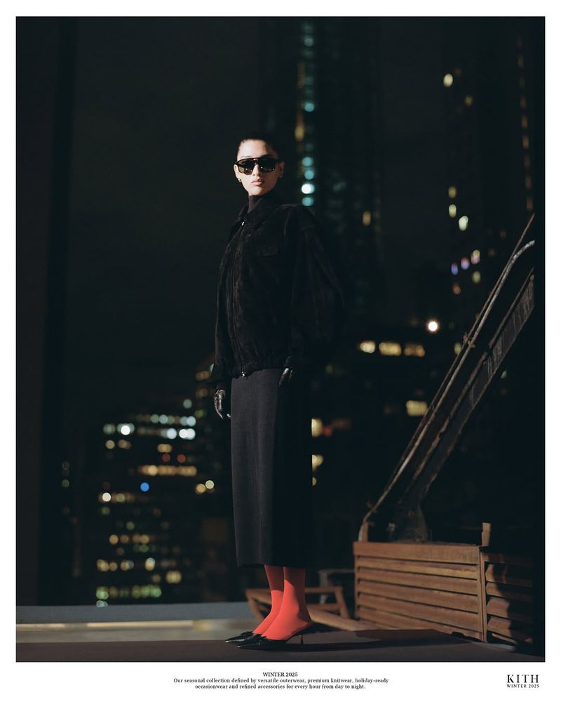 A woman in black clothing stands on a balcony at night in a fashion campaign photo, city lights are out of focus in the background.