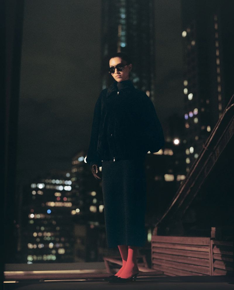 Fashionable woman wearing sunglasses, a dark jacket and skirt, and red tights poses at night with blurred city lights in the background.