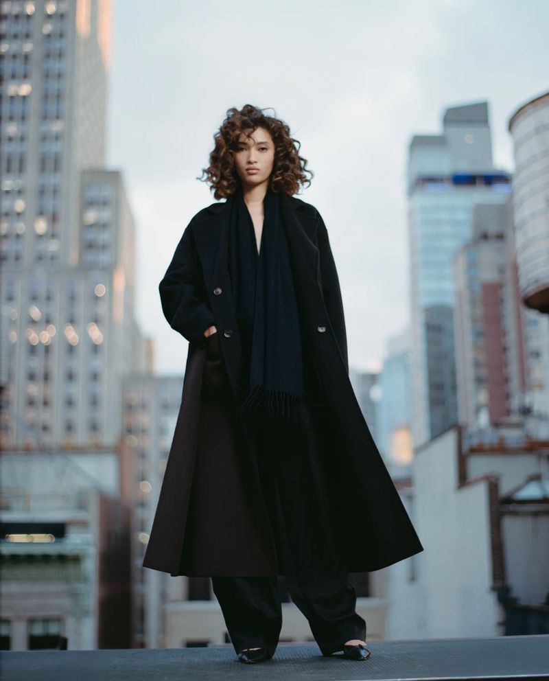 Adult female with curly hair in a black coat and trousers poses against a cityscape backdrop, looking stylish and confident.