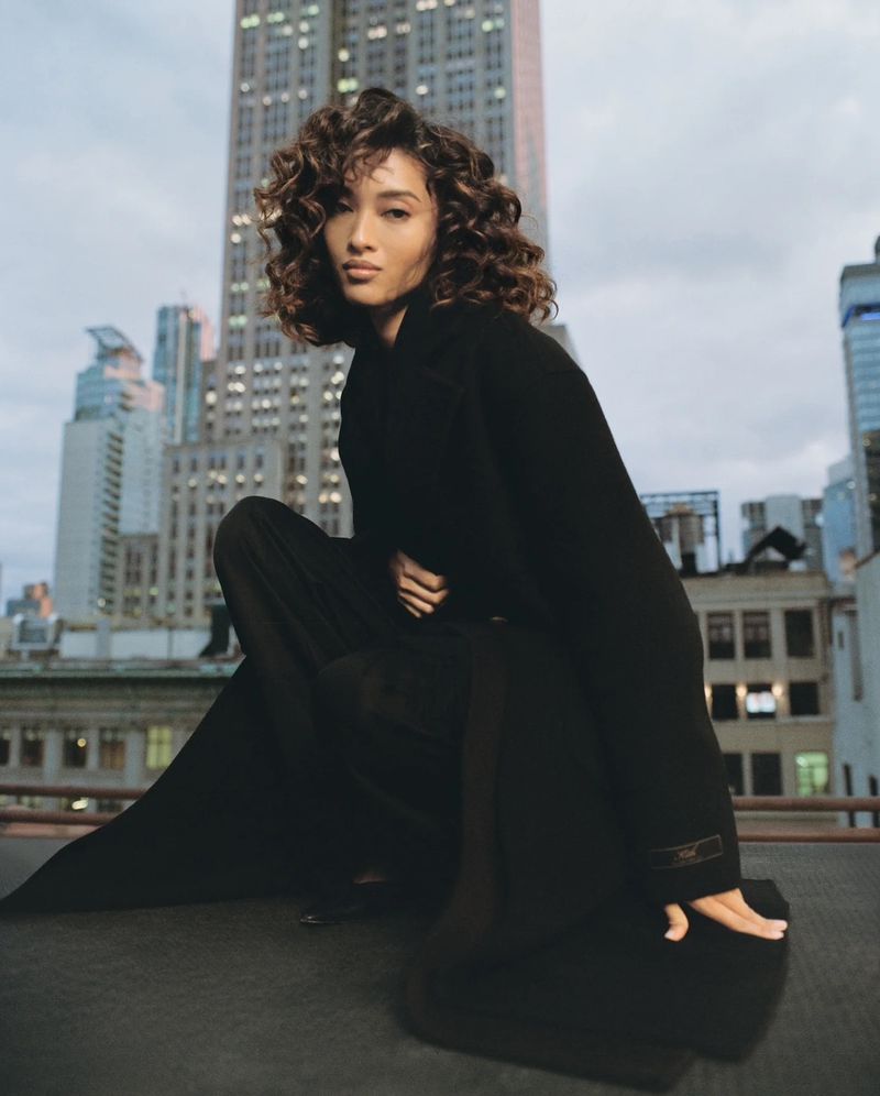 Elegant woman in a black coat poses in an urban setting with city buildings in the background, creating a striking fashion portrait.