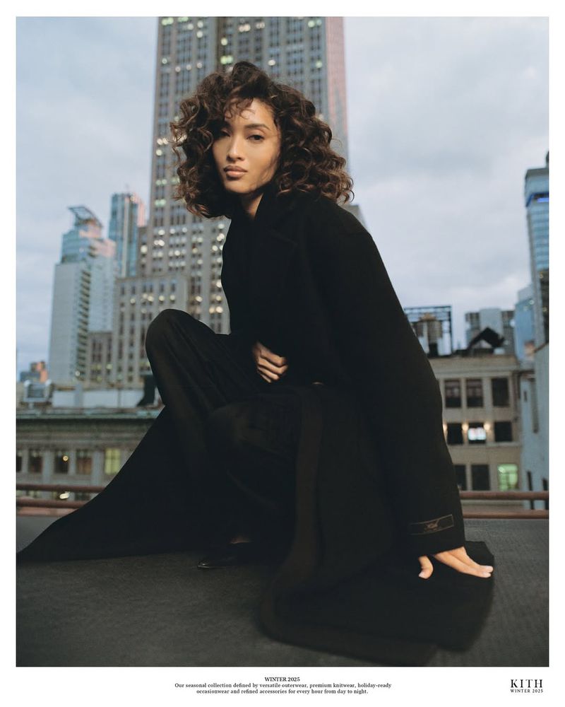 A woman with curly hair, wearing a black coat, poses on a rooftop with skyscrapers in the background. Moody, overcast sky. Fashion editorial.