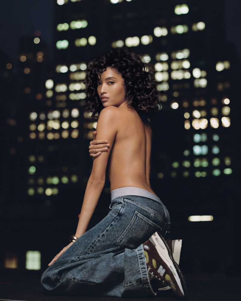 Woman with curly hair poses in jeans and athletic shoes on rooftop at night, city buildings with lights in background, fashion photography.