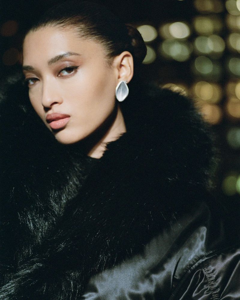 Portrait of a woman with dark hair and fair skin wearing silver earrings and a black jacket against a bokeh background.