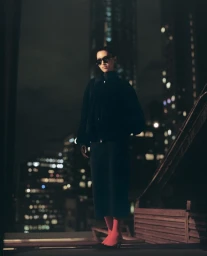 Model Aqua Parios in dark sunglasses, a black jacket, midi skirt, and orange tights standing on a city rooftop at night.