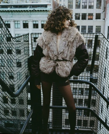 Model with voluminous curly hair wearing a fluffy faux fur vest on a rooftop staircase with city architecture in the background.