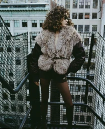 Model with voluminous curly hair wearing a fluffy faux fur vest on a rooftop staircase with city architecture in the background.